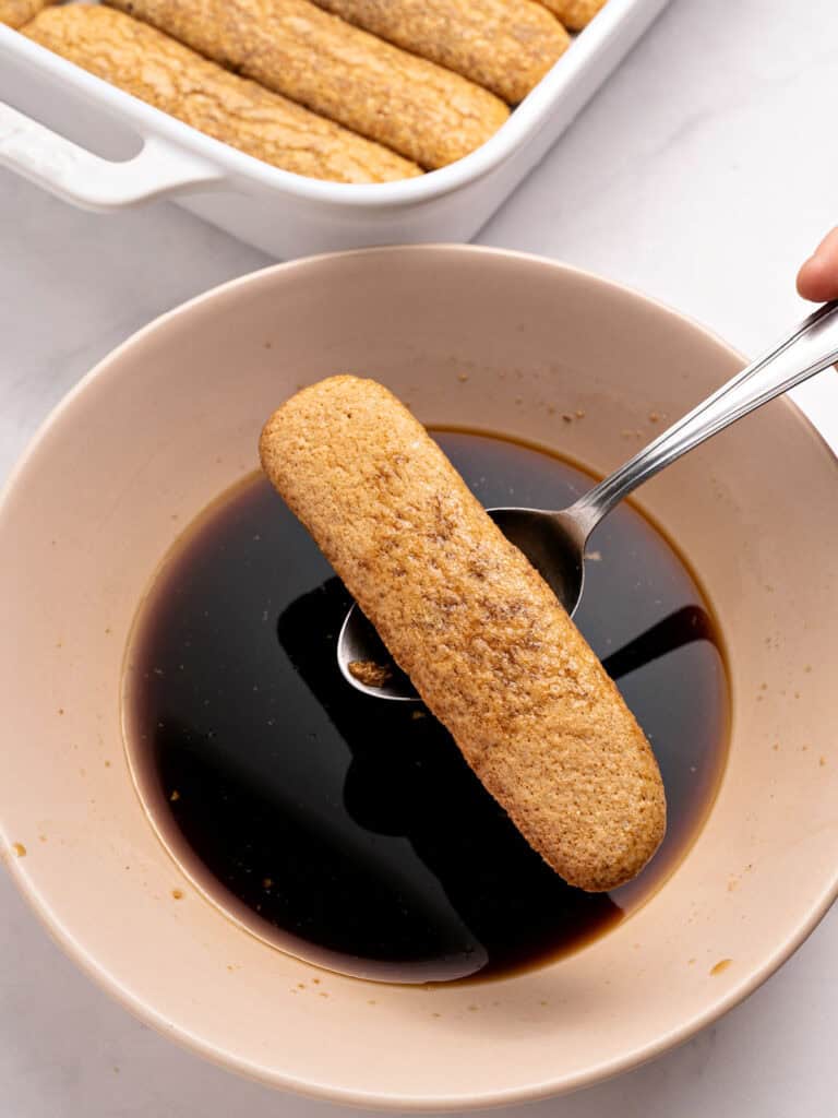 Lifting soaked ladyfinger from pink bowl with coffee.