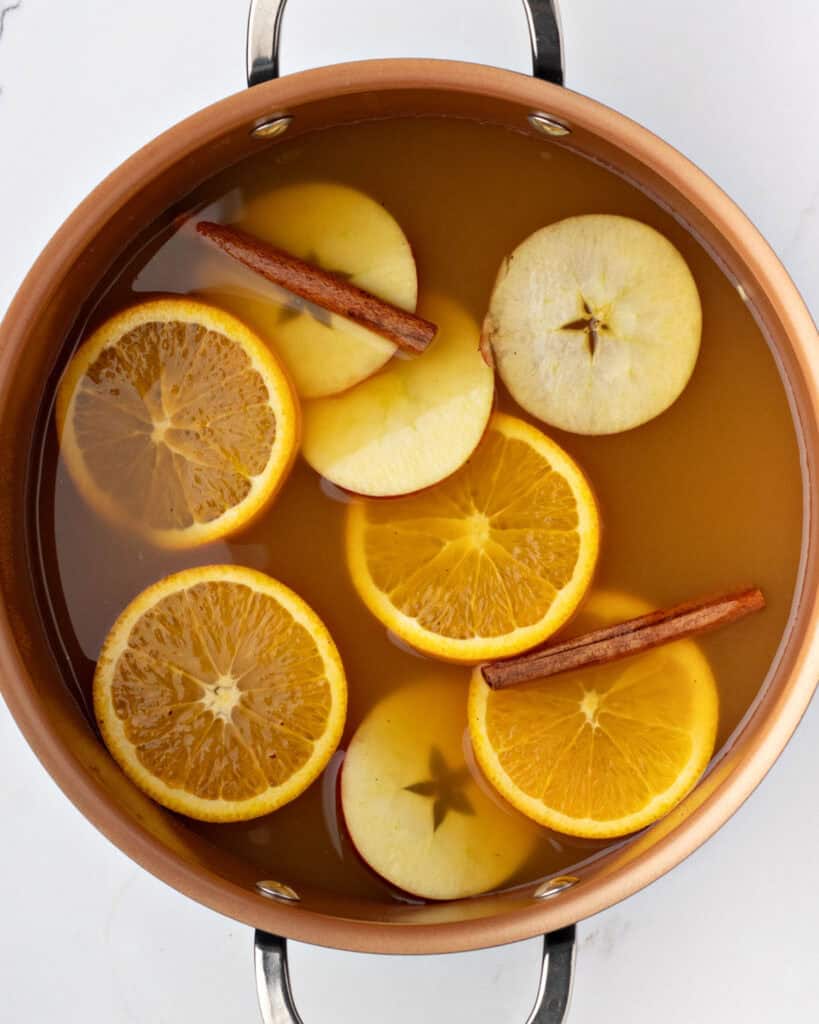 Copper pot with apple cider, cinnamon sticks, orange and apple rounds. White background.