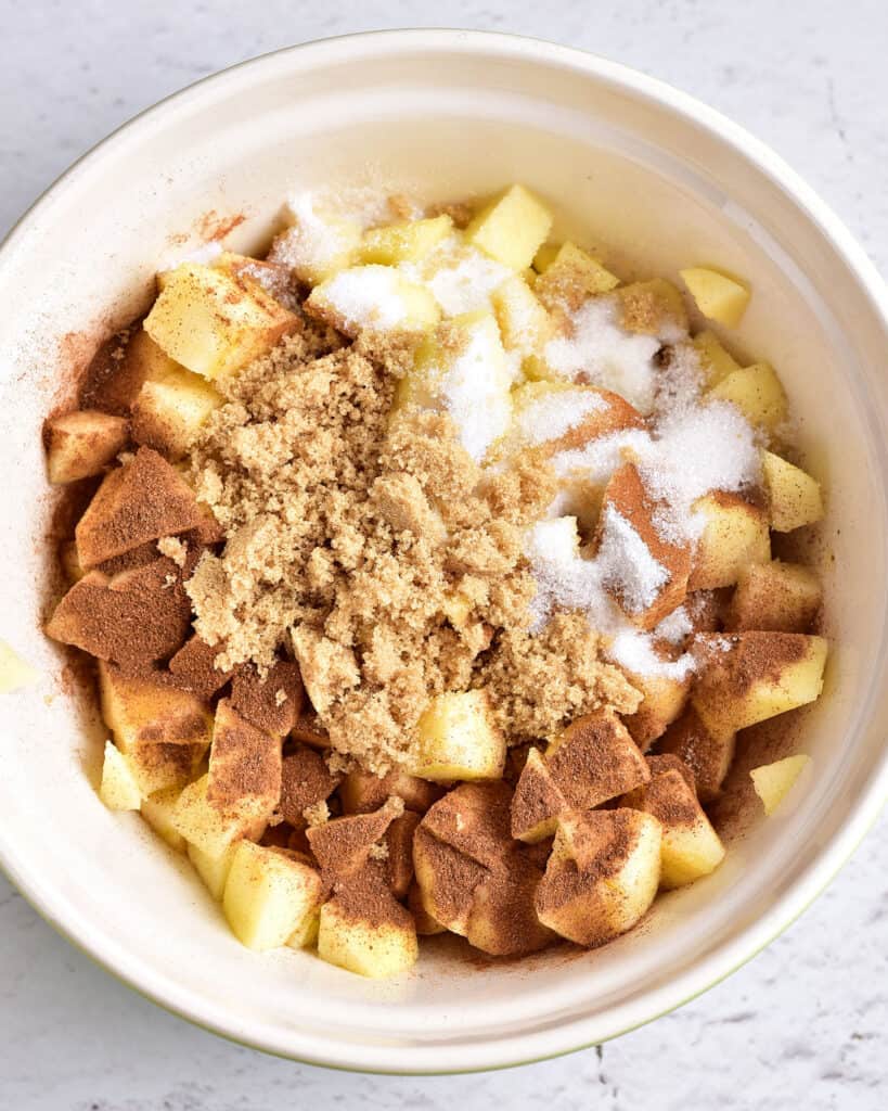 Brown and white sugar, spices and cubed apples in a white bowl.