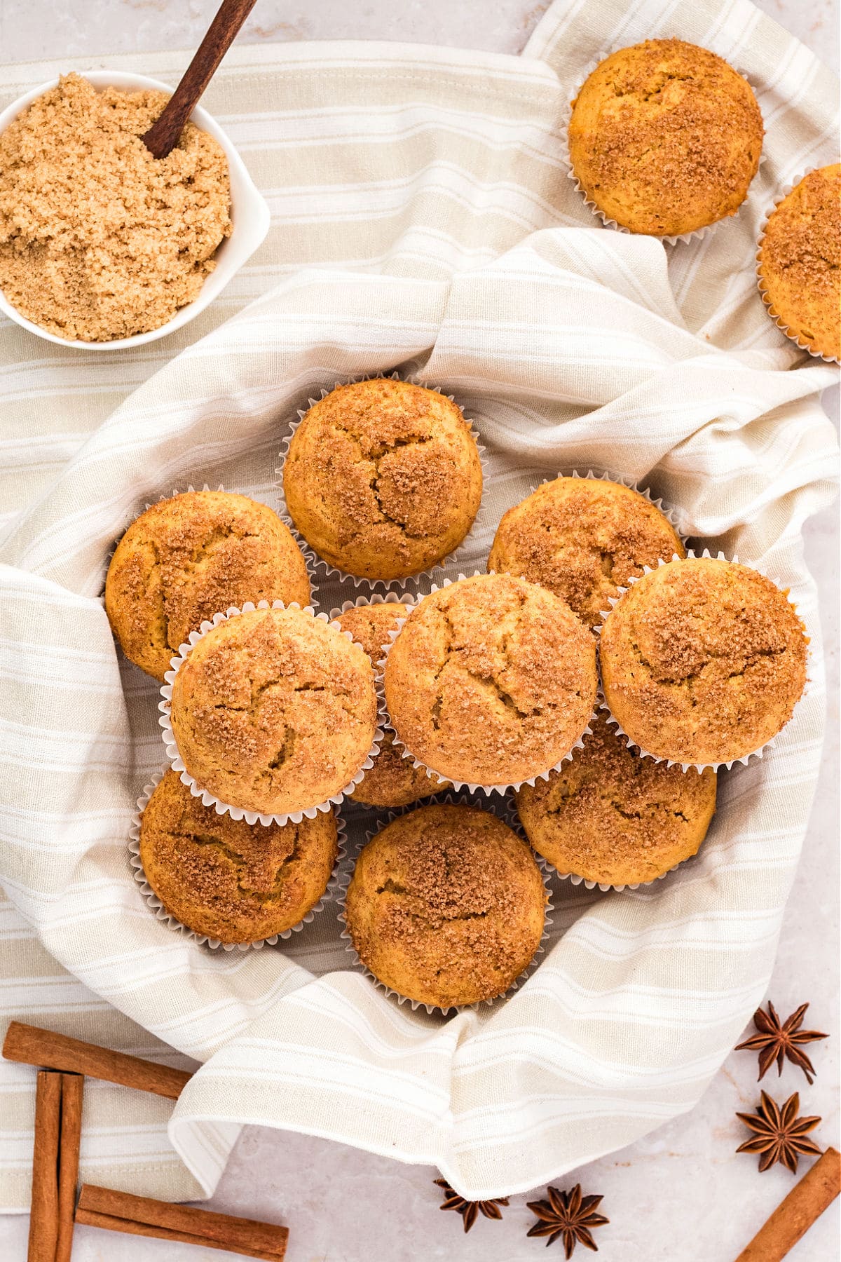 Beige striped cloth with pile of sweet potato muffins. Bowl with brown sugar, cinnamon sticks.