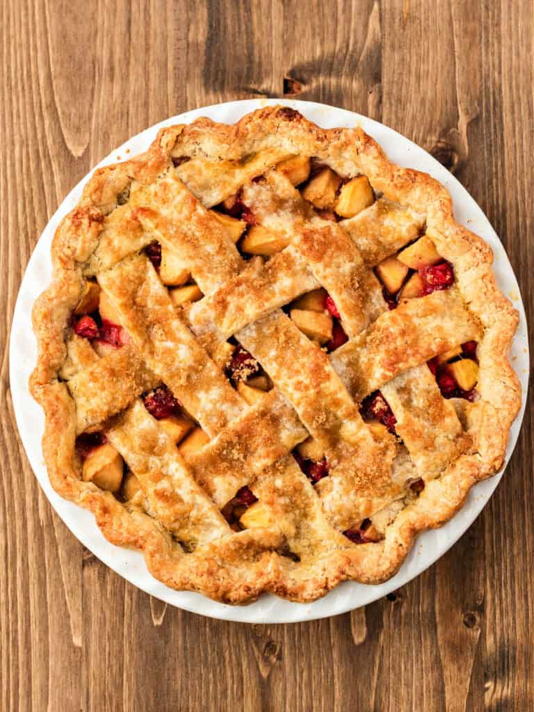 Baked whole apple cran lattice pie in a white dish on a wooden surface.