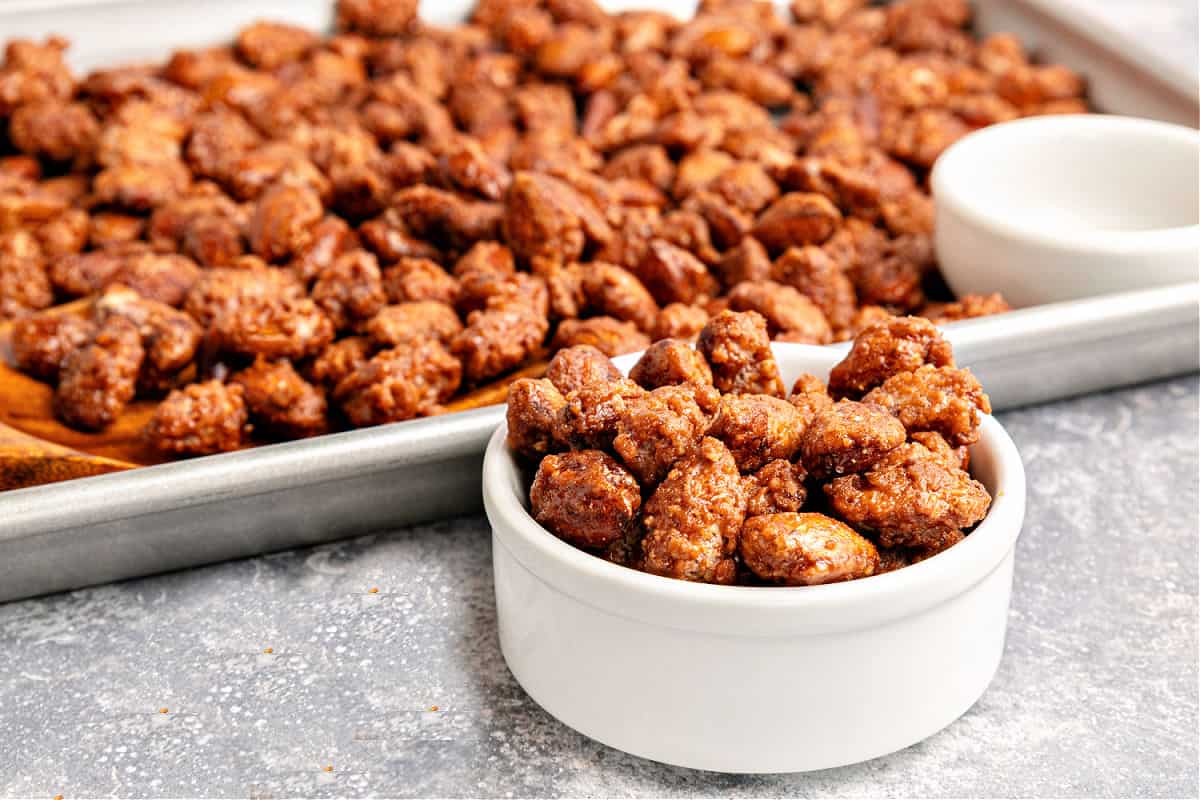 White bowl and baking sheet with candied almonds. Gray surface.