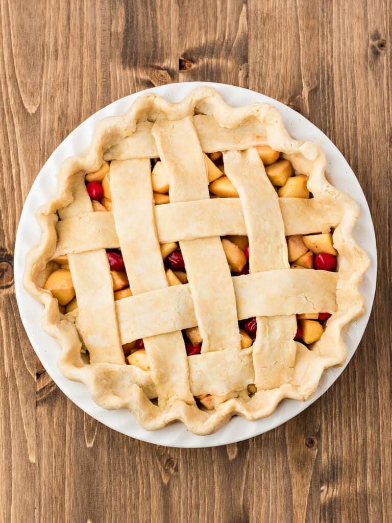 Top view on a wooden surface of white dish with lattice apple cranberry pie before baking.