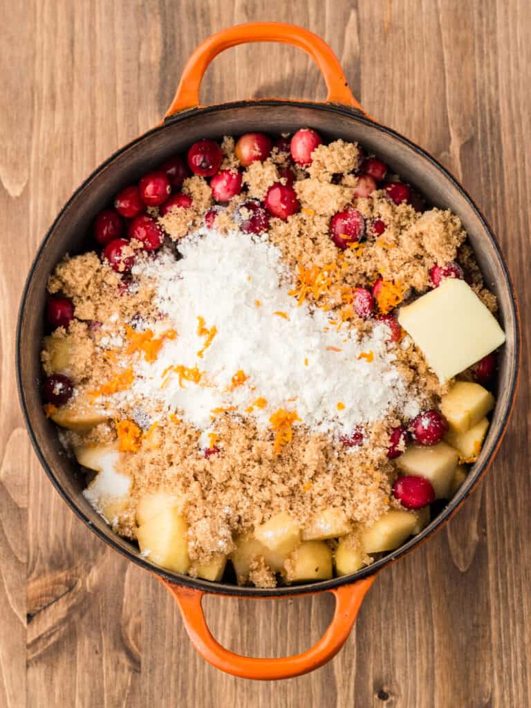 Saucepan with apples, sugars, butter, orange zest and cranberries. Wooden background.