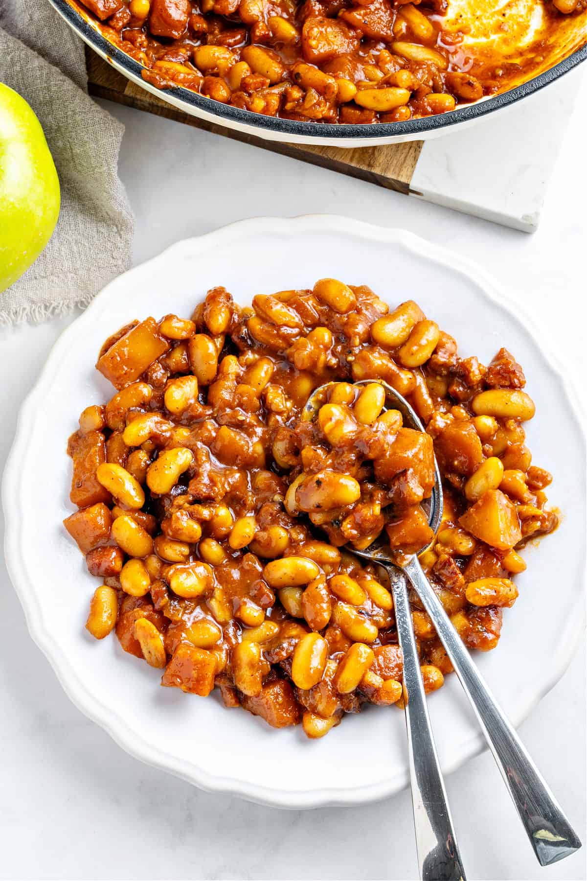 White plate with serving of baked beans and apples. Silver cutlery. White background.