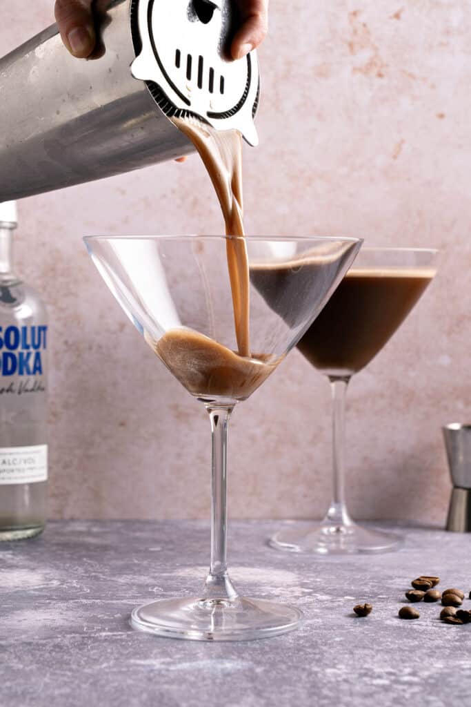 Pouring espresso chocolate martini into stem glasses from a shaker. Reddish purple background.
