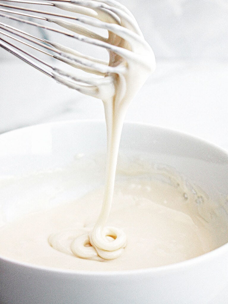Wire whisk dripping powdered sugar glaze. White bowl and background.