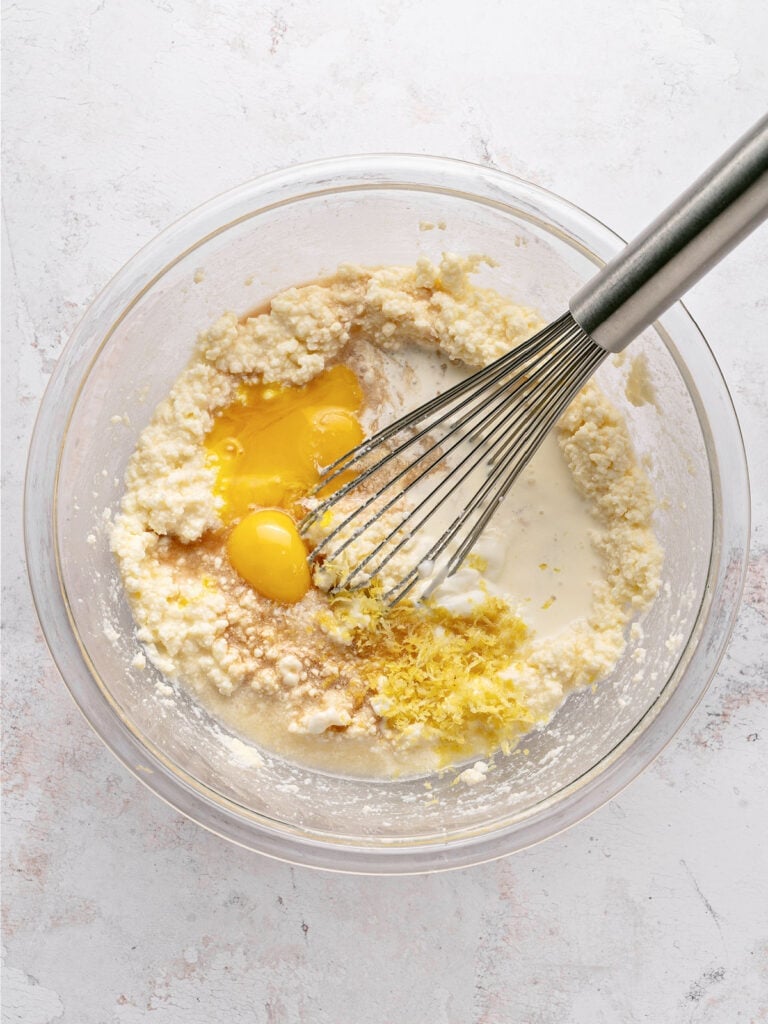 Glass bowl with lemon ricotta pie filling ingredients. Metal whisk. Light gray background. 