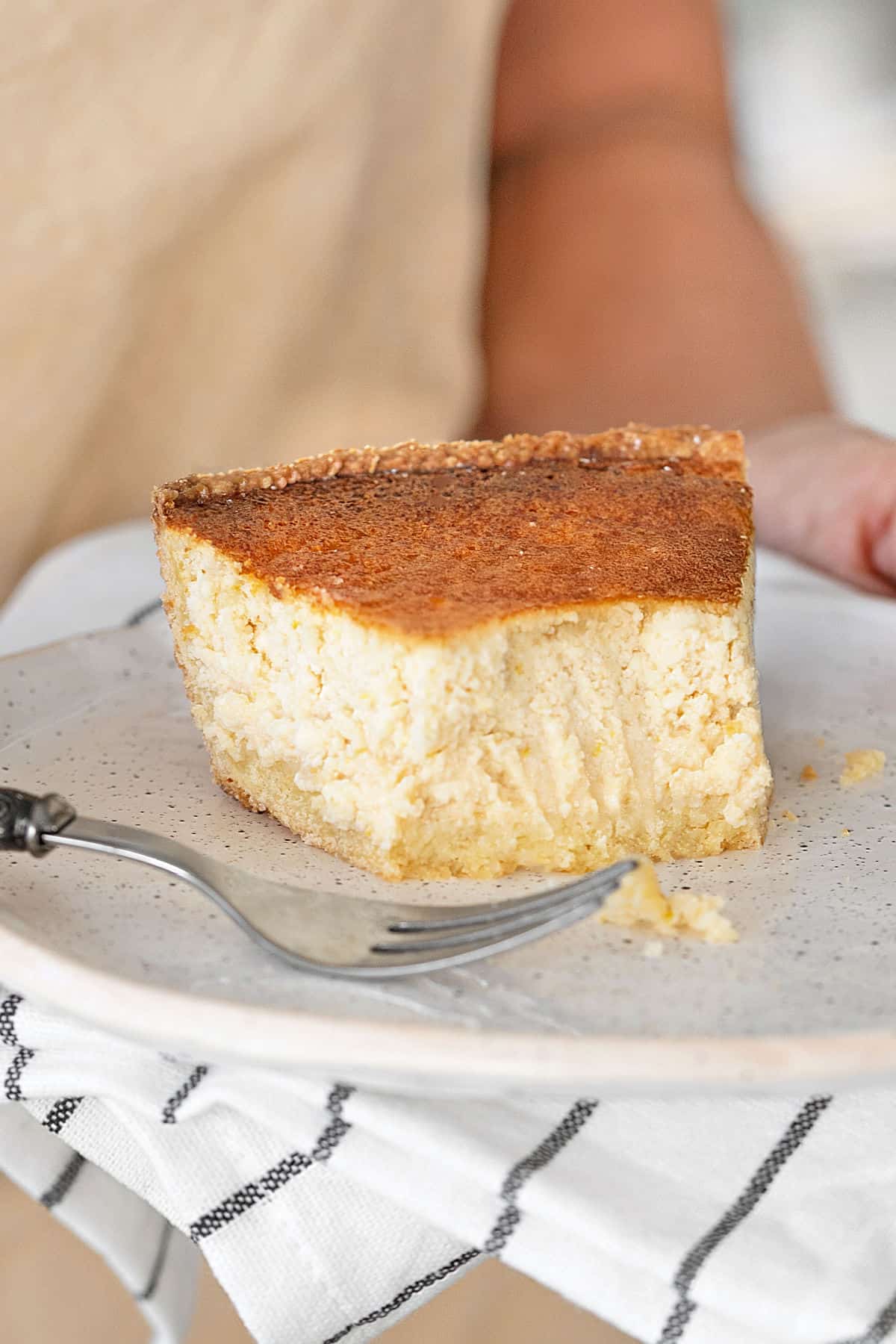 Forked slice of ricotta pie held on a white plate. Gray white striped cloth.