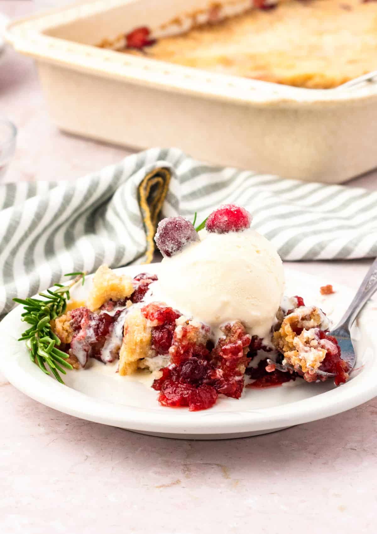 Serving of cranberry cobbler with ice cream on a white plate. Green striped cloth, cream-colored background and dish.