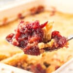 Close up spoon with cranberry cake mix cobbler above dish with dessert.