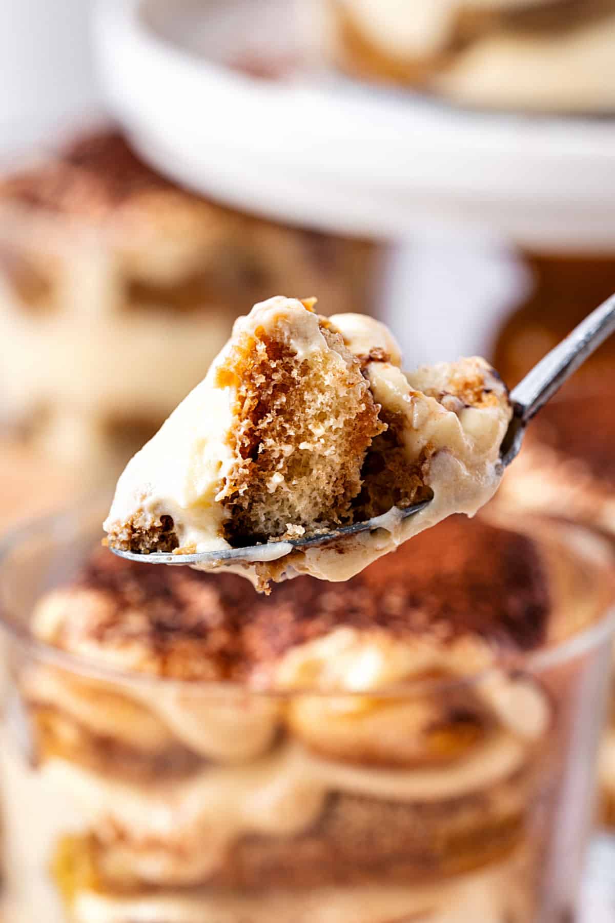 Bite of tiramisu on a spoon. More cups and white stand in the background.