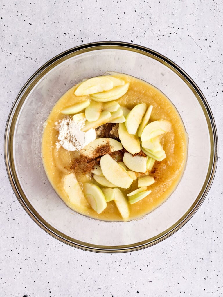Glass bowl with apple wedges, applesauce, flour and spices. Light gray background.
