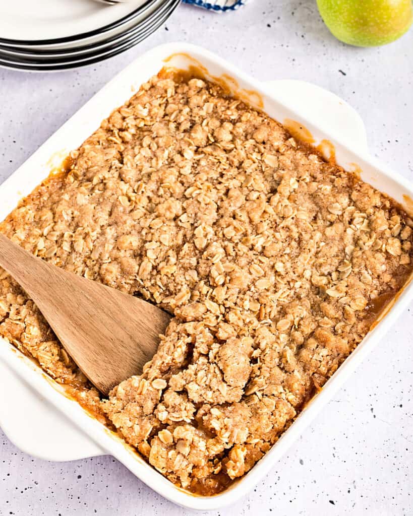 Baked applesauce crisp in a white dish. Wooden spoon inside. Light gray background.