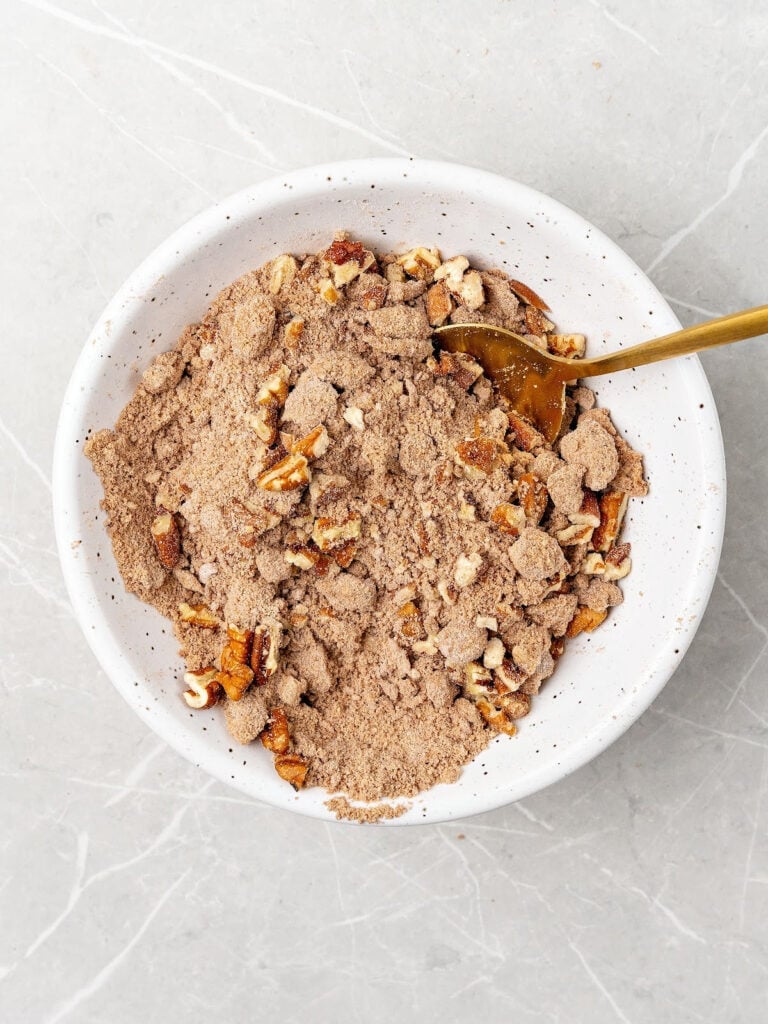 Streusel mixture with pecans in a white bowl. Gold spoon. LIght gray background.