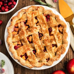 Lattice apple cranberry pie in a white dish. Dark wood surface, gold cake server.