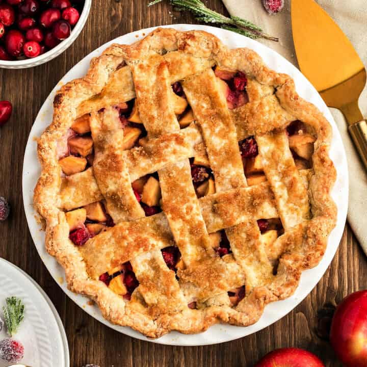 Lattice apple cranberry pie in a white dish. Dark wood surface, gold cake server.