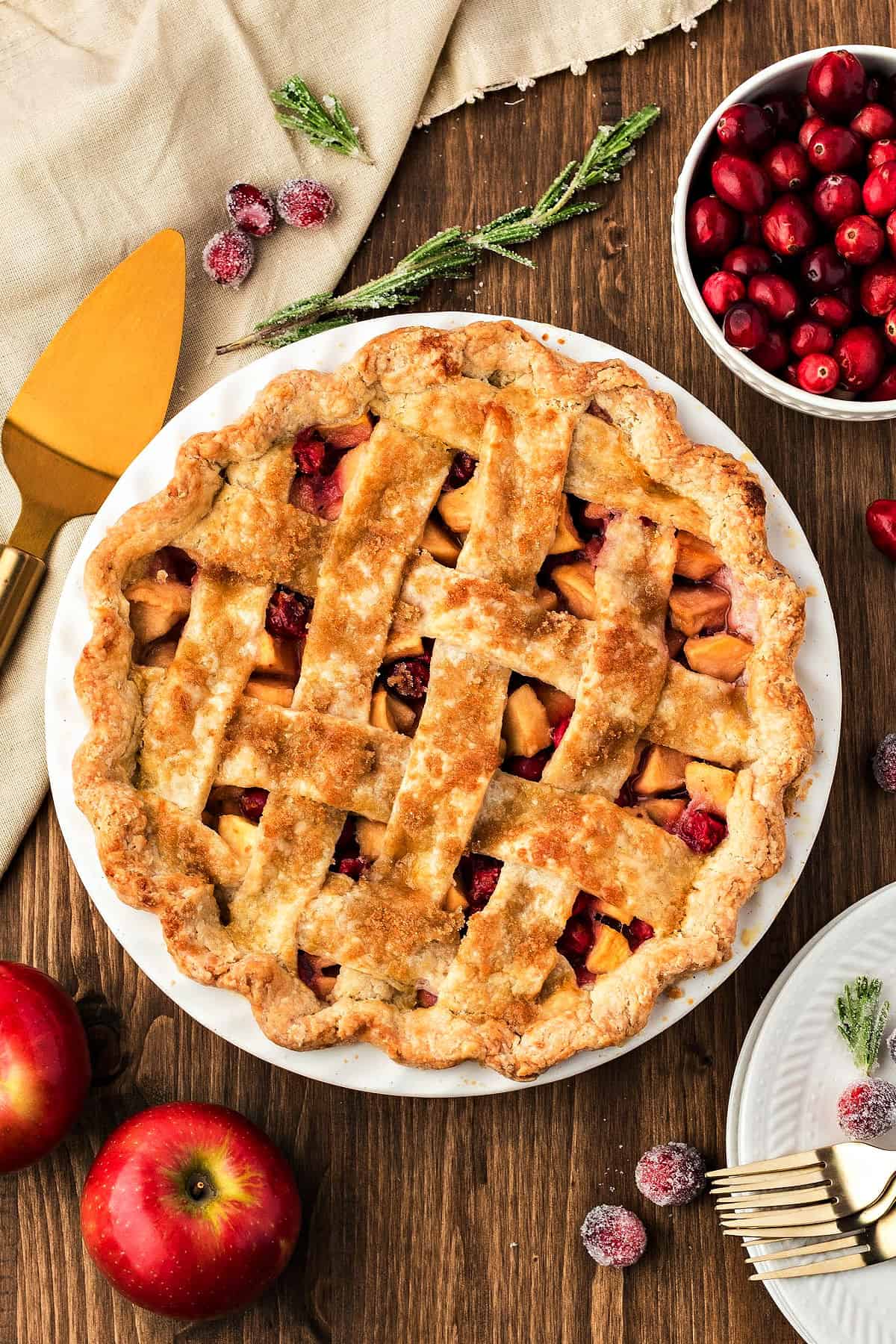 Whole lattice apple cranberry pie in a white dish. Dark wood surface, fresh apples and berries, beige cloth.