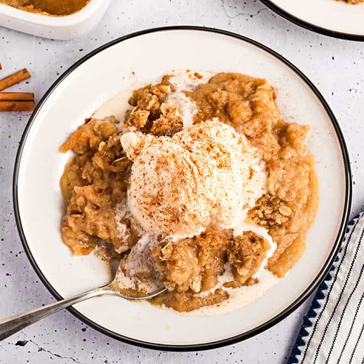 Close up serving of ice cream topped serving of applesauce crisp on a black-rimmed white enameled plate.
