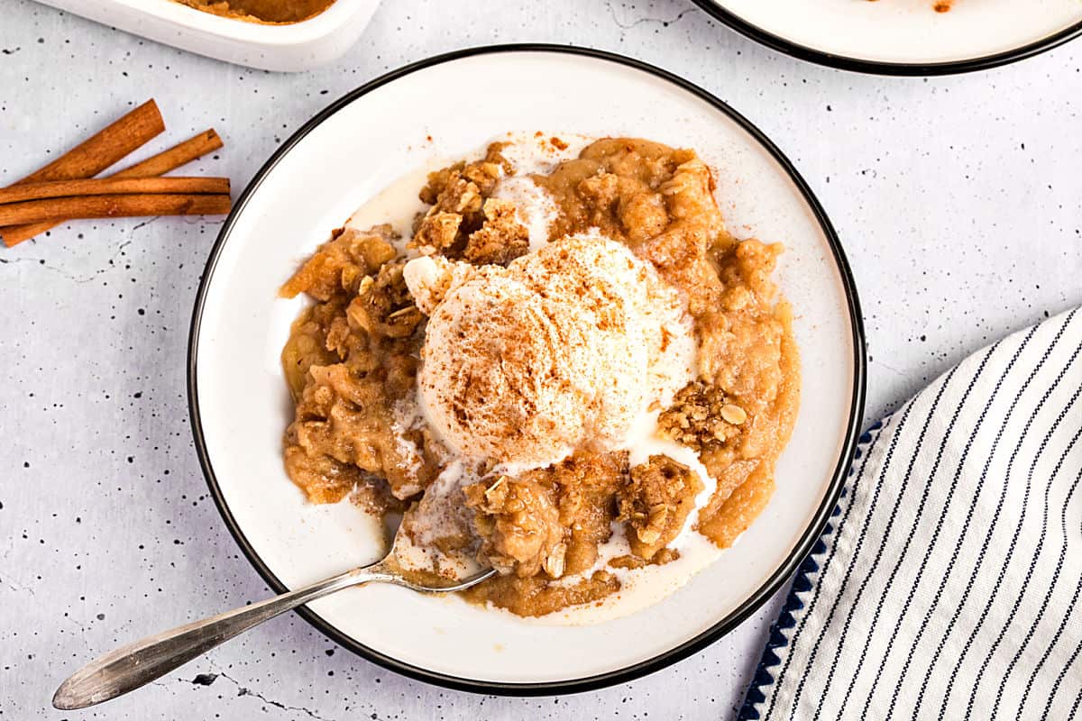 Horizontal image of ice cream topped applesauce crisp on a black-rimmed plate. Light gray background. 