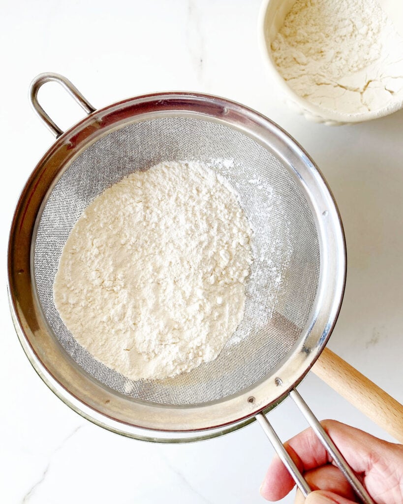 Sifting flour mixture. White surface, white bowl with flour.