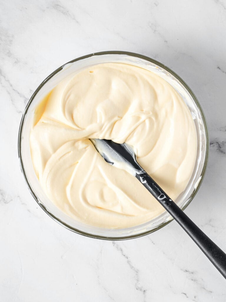Creamy mascarpone mixture for tiramisu. Glass bowl, gray spatula, white background.