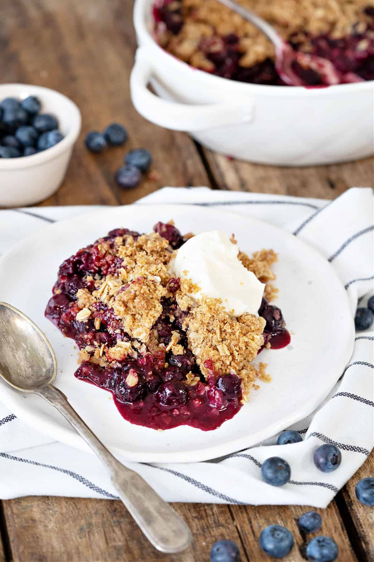 Blue striped kitchen towel and wooden surface with white plate of blueberry crisp. Silver spoon.