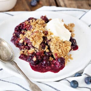 Striped cloth with white plate of blueberry oat crisp with cream. Silver spoon.