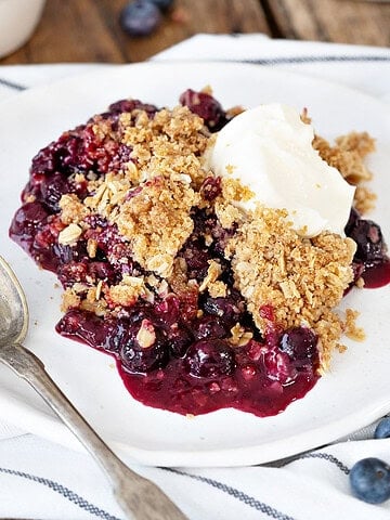 Striped cloth with white plate of blueberry oat crisp with cream. Silver spoon.