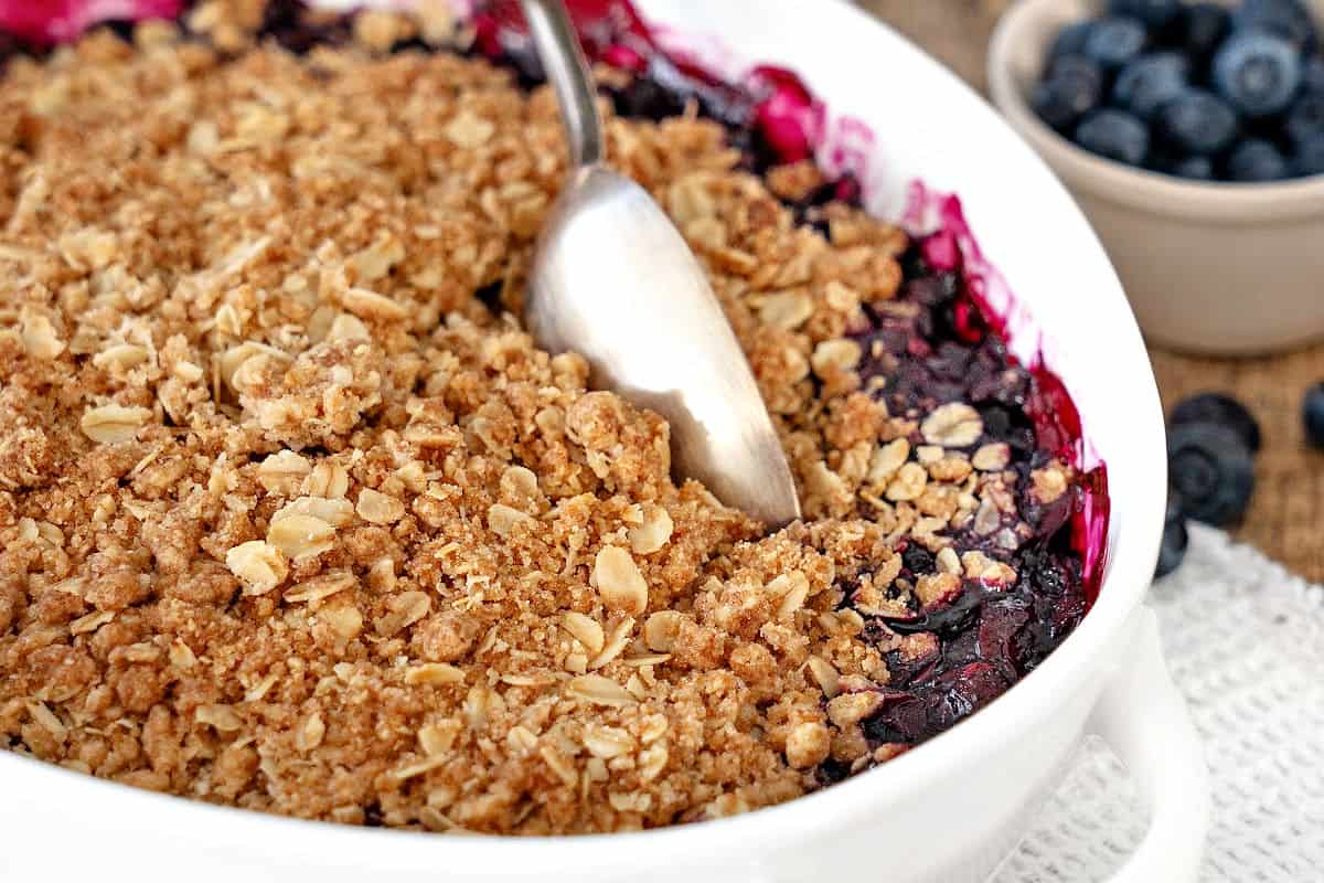 Close up silver spoon scooping blueberry crumble in a white dish.