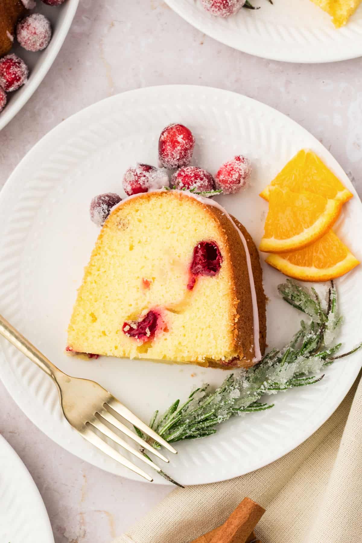 Slice of cranberry pound cake on a white plate. Gold fork, sugared cranberries, orange and rosemary.