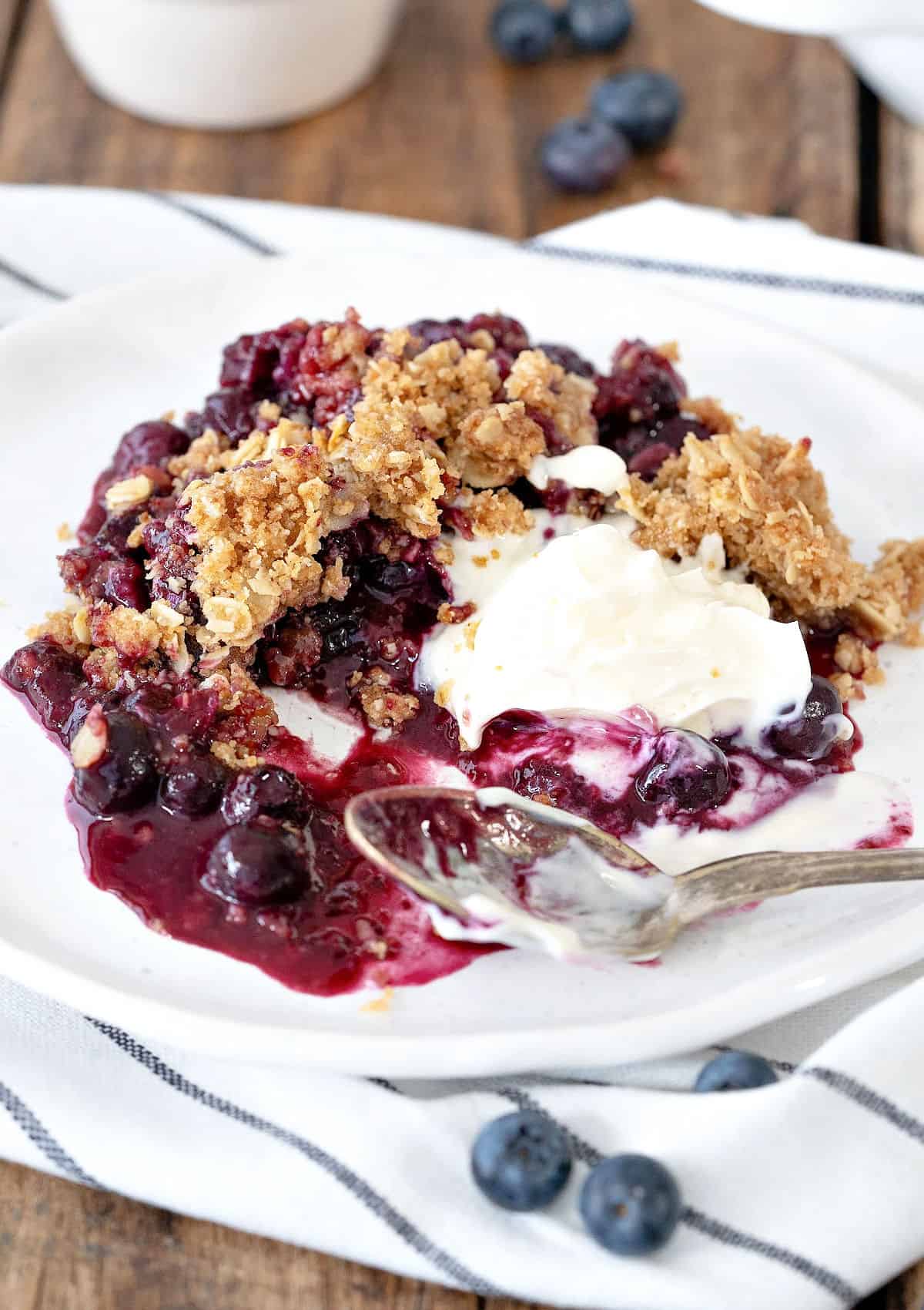 Blueberry oat crumble serving with cream on a white plate. Silver spoon, striped towel.