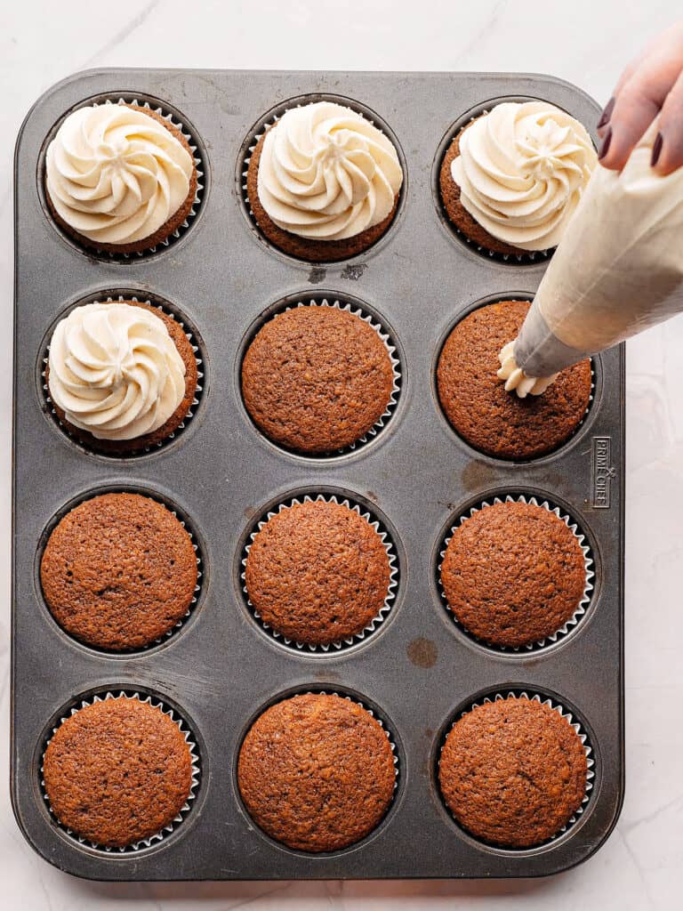 Piping spiced frosting on gingerbread cupcakes in a dark metal pan.