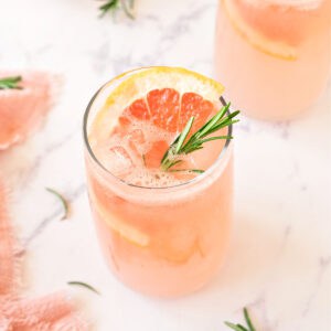 Tall glass with grapefruit cocktail. Rosemary sprig, grapefruit slice, white marble.