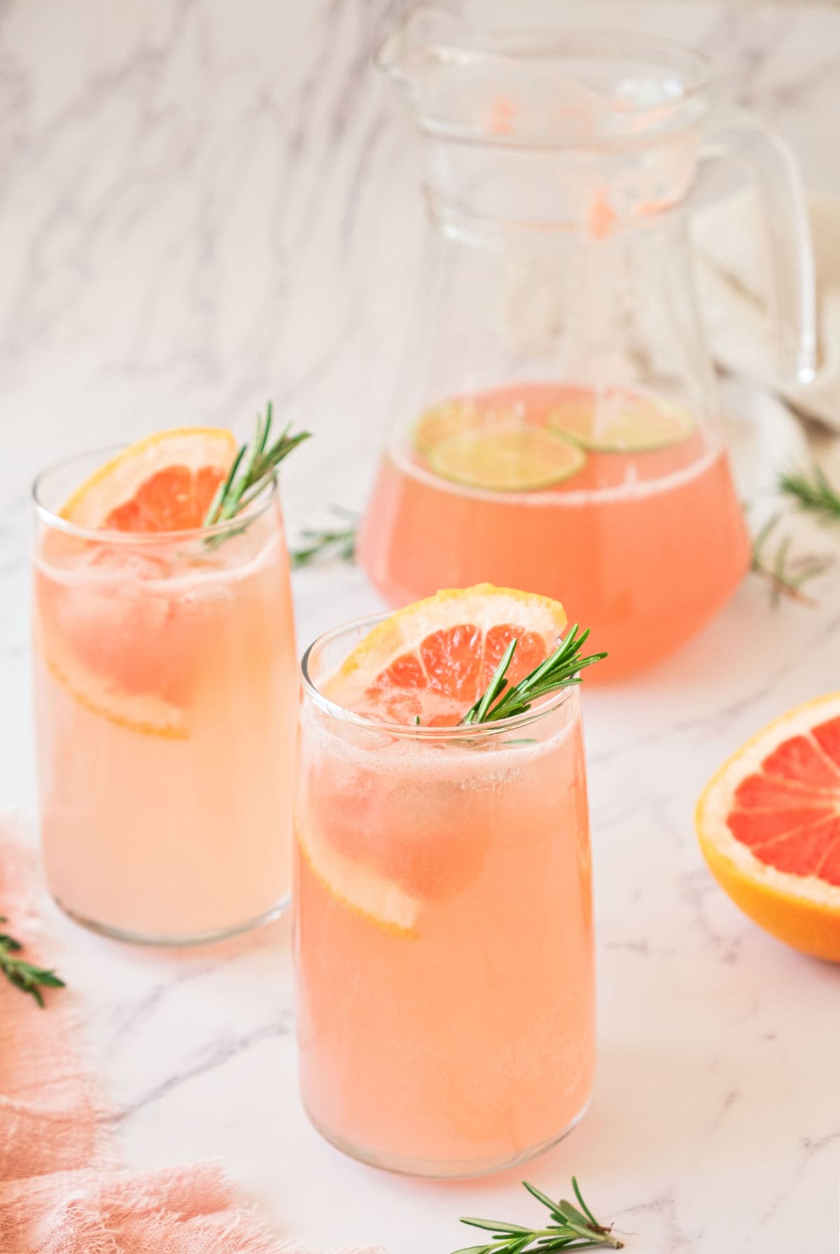 Pitcher and tall glasses of grapefruit drink. Rosemary sprigs, grapefruit slices, marble background.