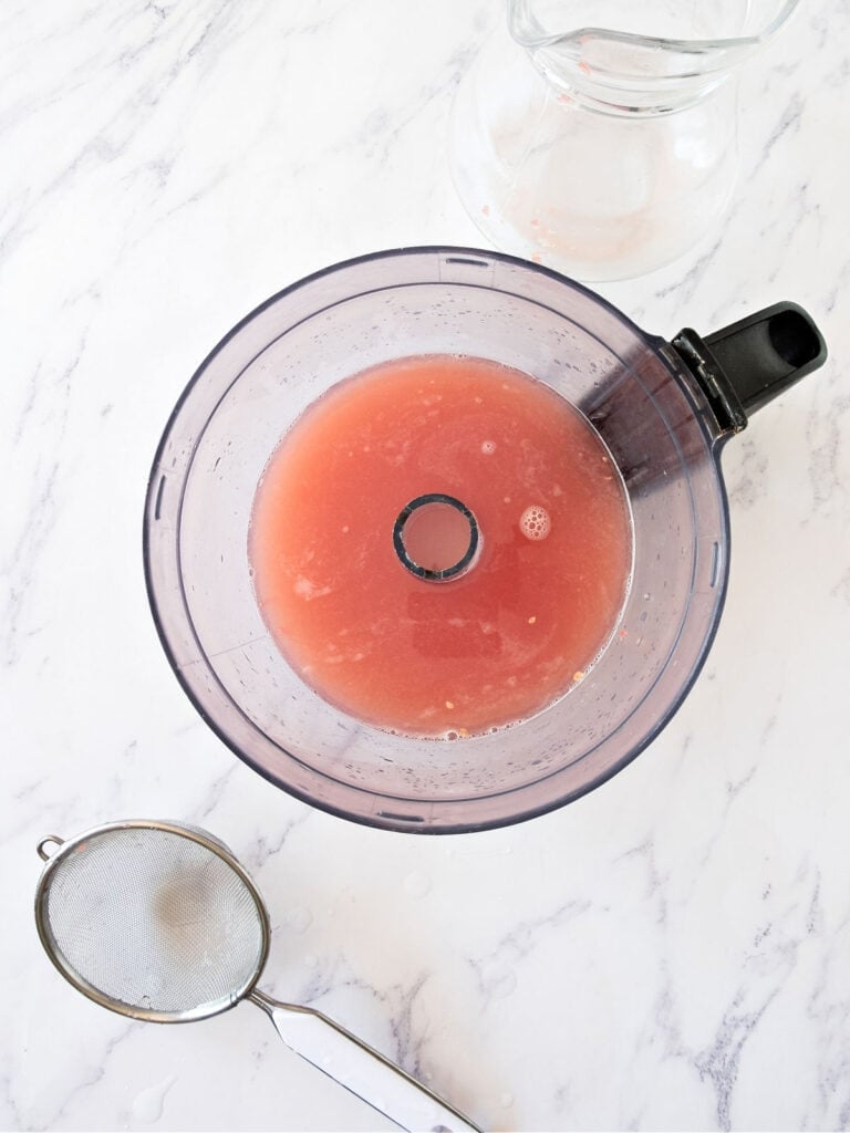 Food processor bowl with grapefruit juice. Colander, white marble surface.