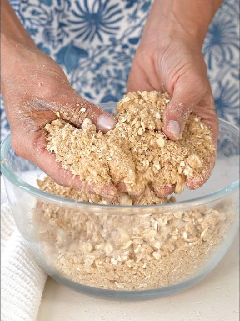 Hands lifting oat crumble mixture from a glass bowl.