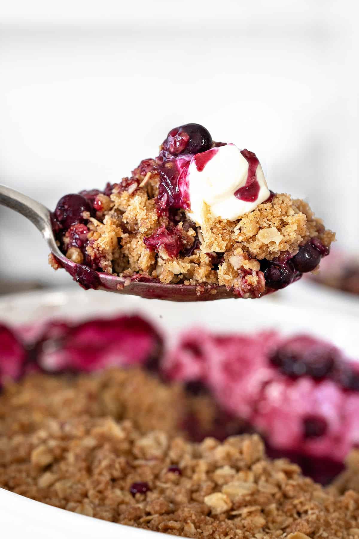 Spoon with cream-topped blueberry crisp lifted over white baking dish.