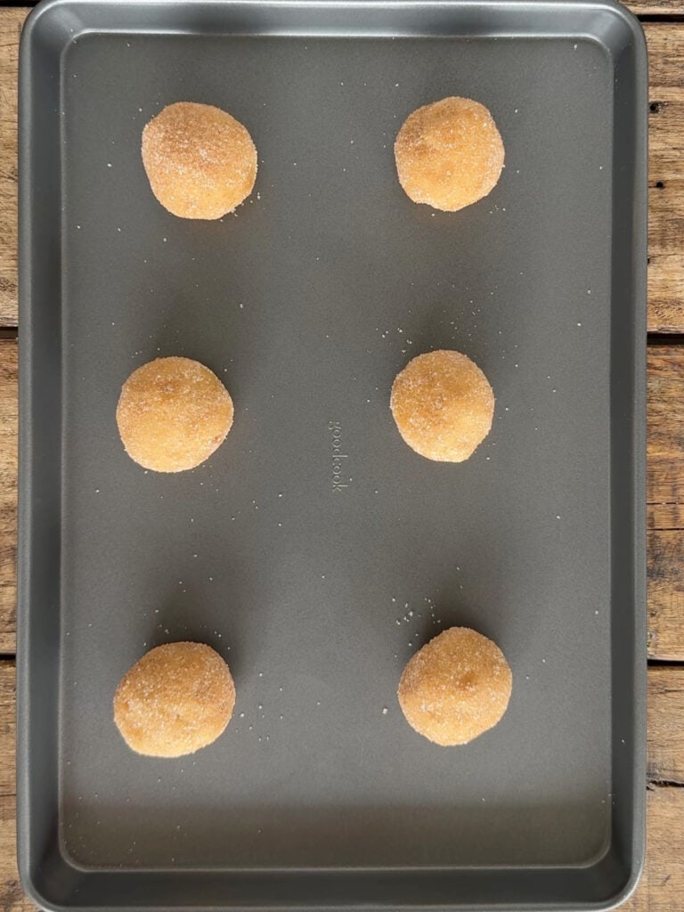 Dark metal baking sheet with six cinnamon sugar coated cookie dough balls.