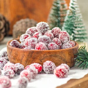Bunch of sugared cranberries in a wooden bowl and white paper. Christmas decor in the background.