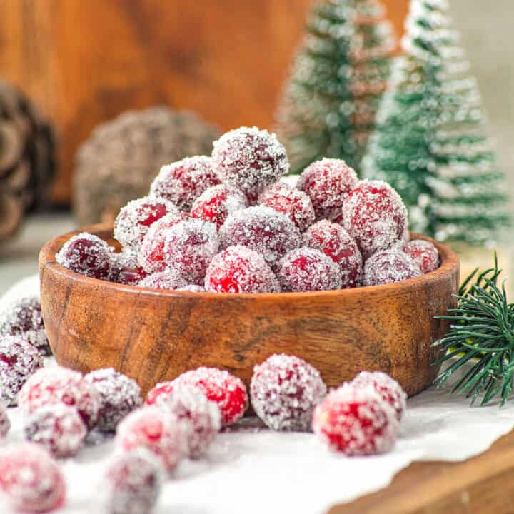 Bunch of sugared cranberries in a wooden bowl and white paper. Christmas decor in the background.