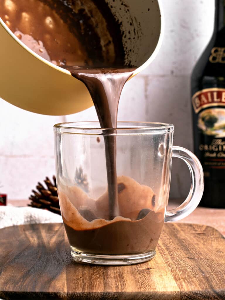 Pouring hot chocolate in a glass mug on a wooden board.