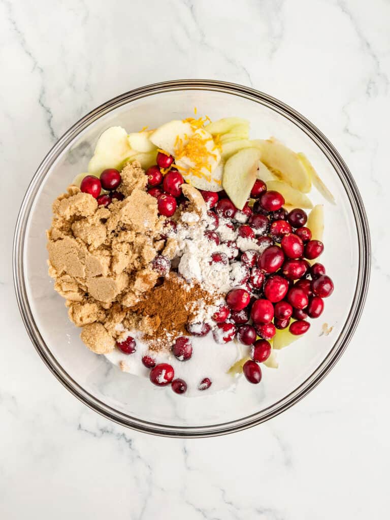 Glass bowl with ingredients for apple cranberry pie filling. White marble background.