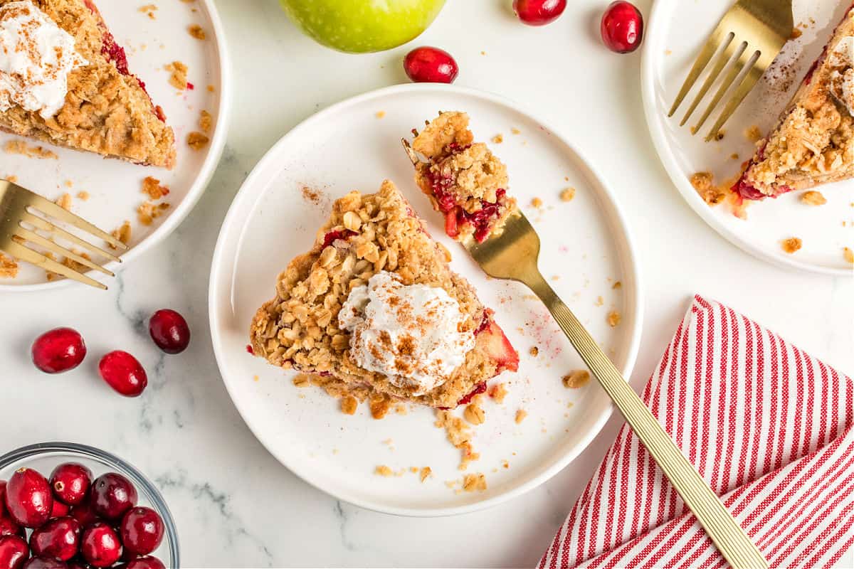 Several white plates with slices of cream-topped apple cranberry crumb pie. Gold forks, striped cloth, white surface.