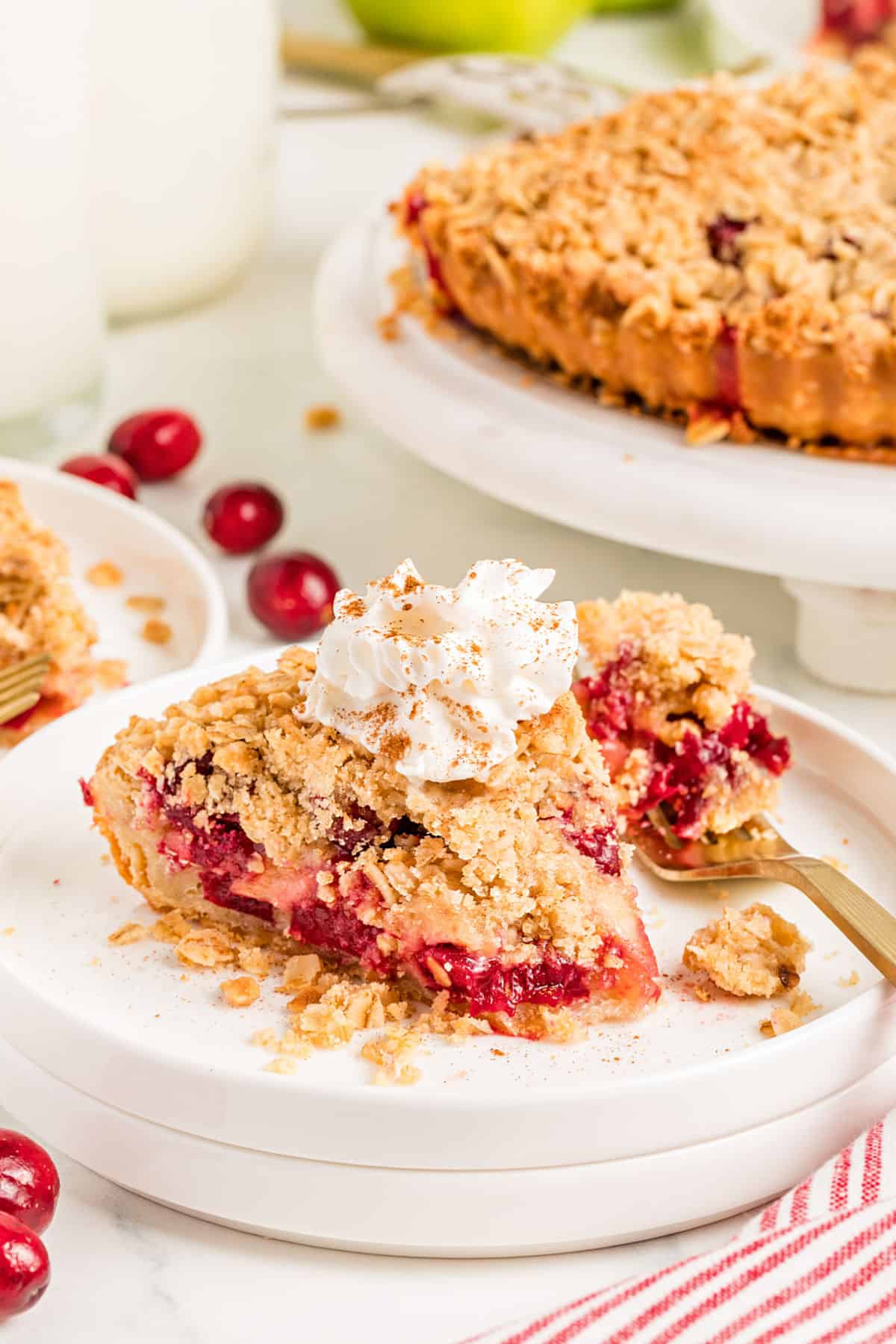 Forked slice of apple cranberry crumble pie on a white plate. Ice cream topped, pie on cake stand.