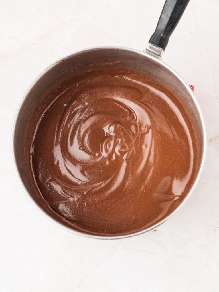 Chocolate custard for frosting in a metal saucepan on a white surface.