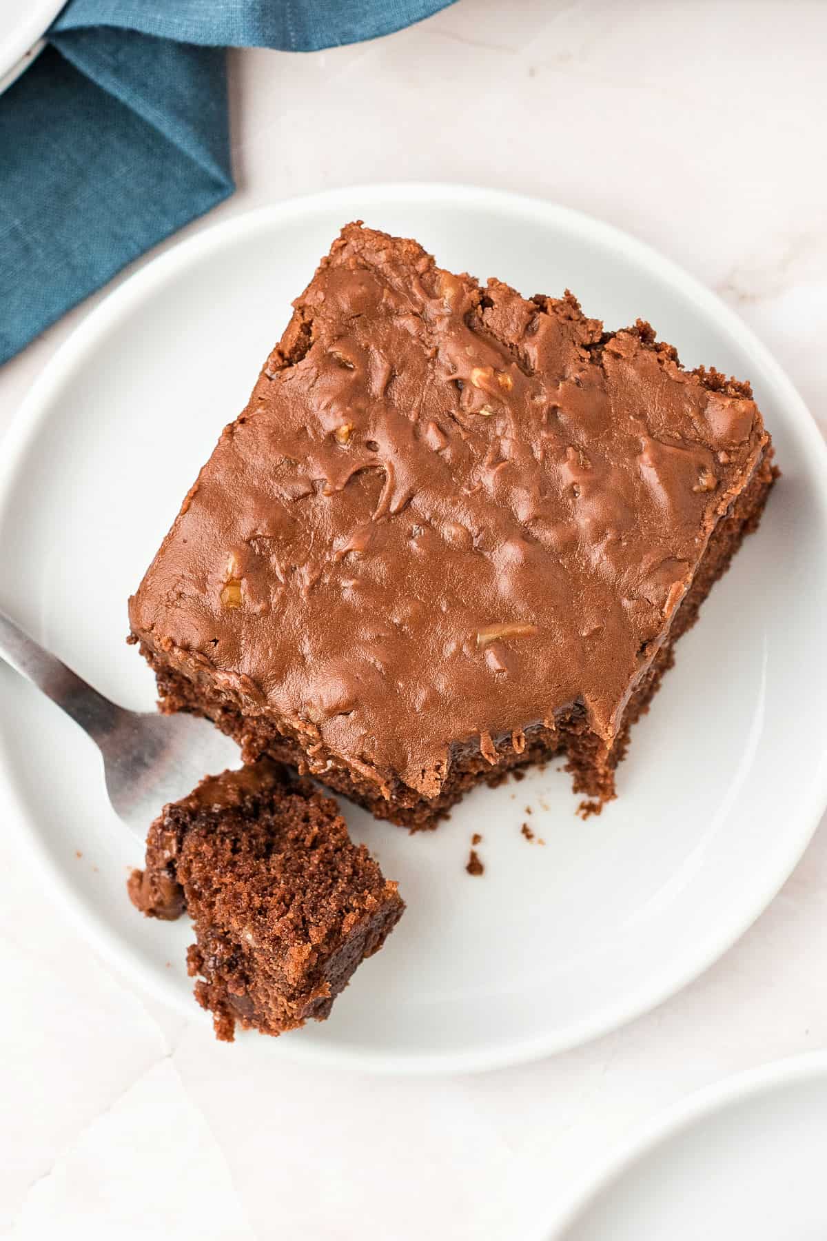 White plate with square of chocolate frosted cake. Silver fork. White surface.