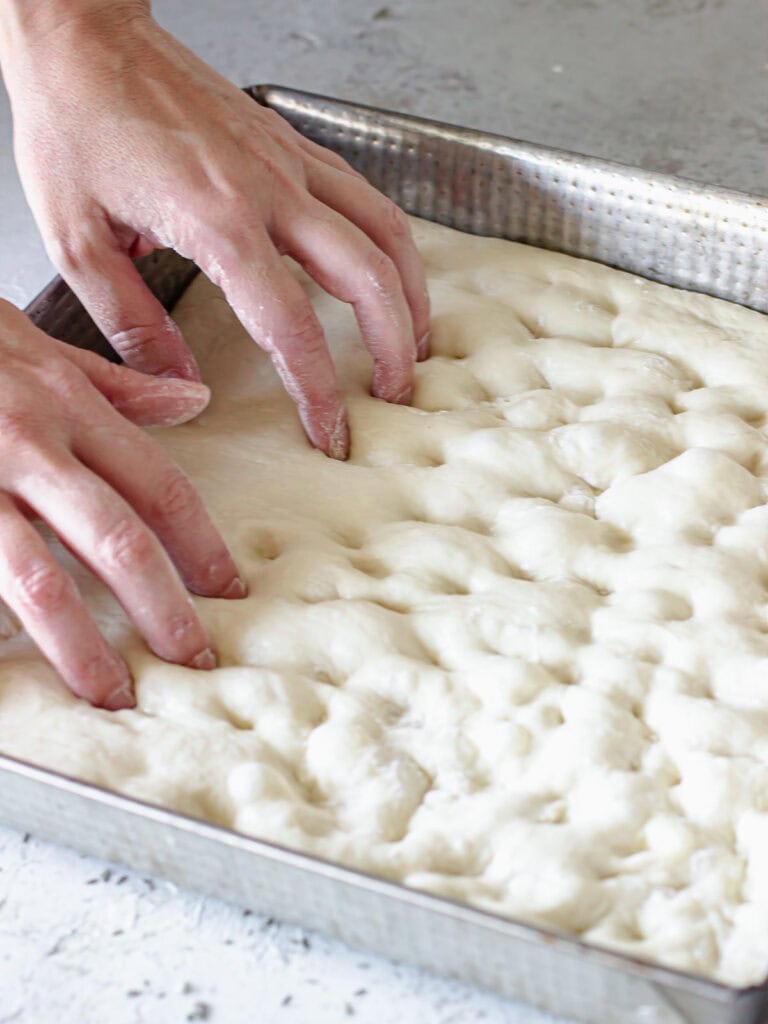 Hands dimpling focaccia dough in a square light metal baking pan. 