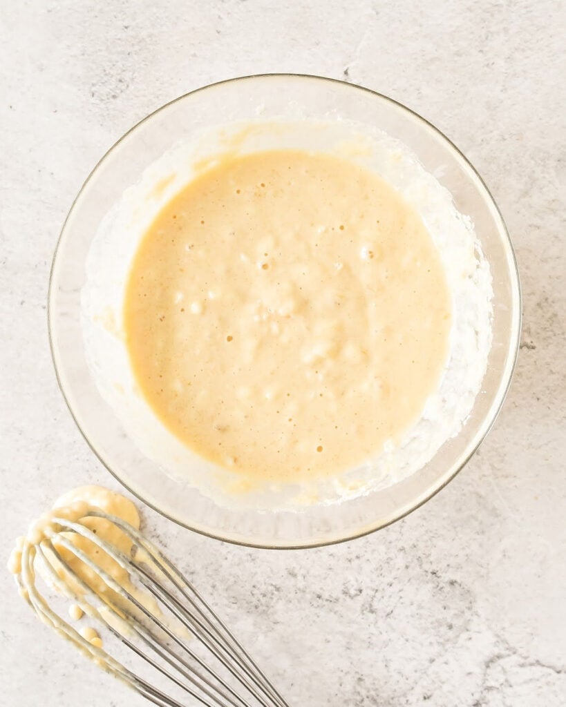 Pancake batter in a glass bowl. Metal whisk. Light gray background.