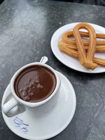 White cup with hot chocolate and plate of churros. Dark surface.